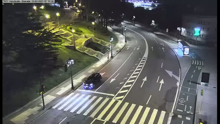 A dark-colored SUV drives across a pedestrian crosswalk on a curving, well-lit road at night.