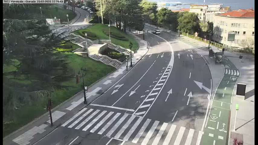 A paved road curves through a tree-lined area with pedestrian crosswalks, a bike lane, and stairs leading up a grassy hillside on a sunny day.