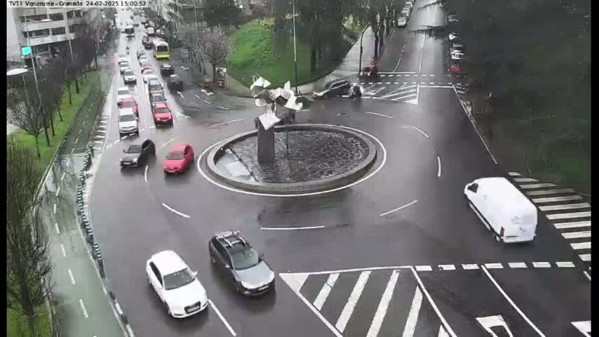 A wet roundabout with several cars navigating it, surrounded by streets with other vehicles and some pedestrian infrastructure.
