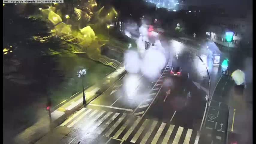 A wet, nighttime street intersection shows vehicles moving through crosswalks and traffic lights under hazy, poorly lit conditions.