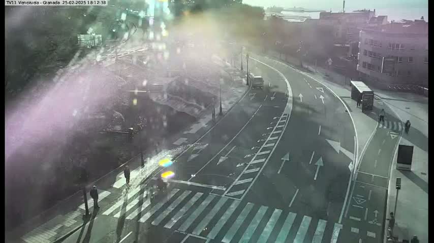 A high-angle, hazy view shows a roadway with a pedestrian crossing, bike lanes, and several people walking, under a bright, possibly sunny sky.