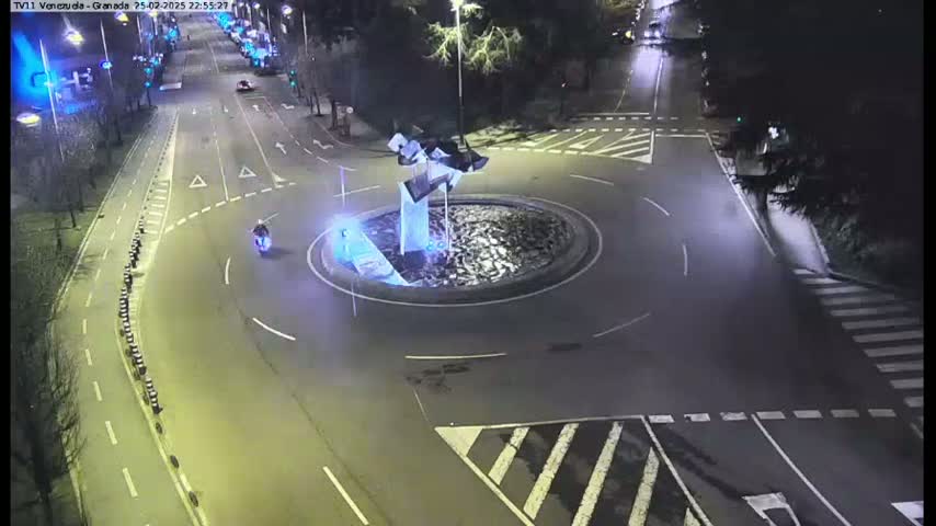 A nighttime, overhead view of a roundabout with a modern sculpture in the center, a motorcycle driving through, and several cars in the distance on a dark roadway.