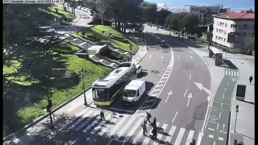 A sunny day reveals a city street scene with a bus and van stopped at a crosswalk, pedestrians crossing, and a curving road leading towards buildings and a body of water in the distance.
