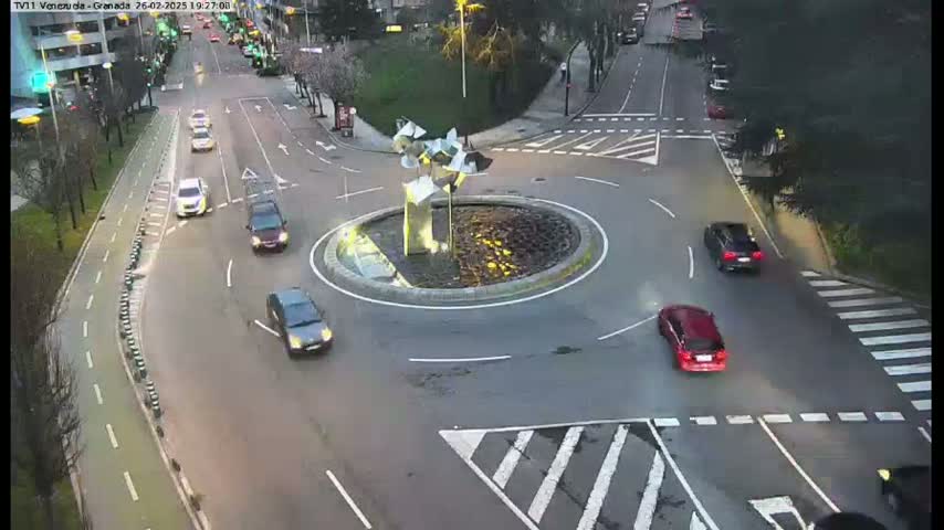 Several cars navigate a roundabout in a city setting at dusk or early evening.