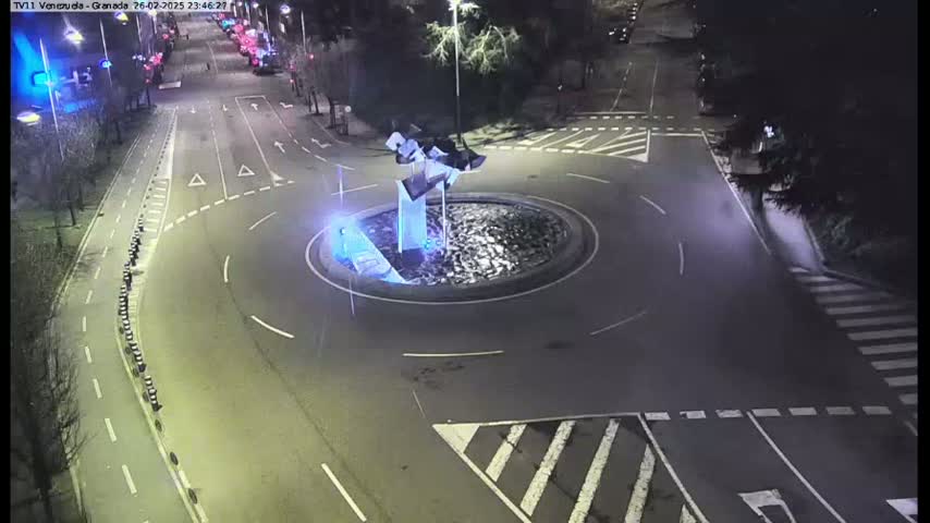 A nighttime aerial view of a roundabout with a modern sculpture in the center, surrounded by roads and pedestrian crossings.