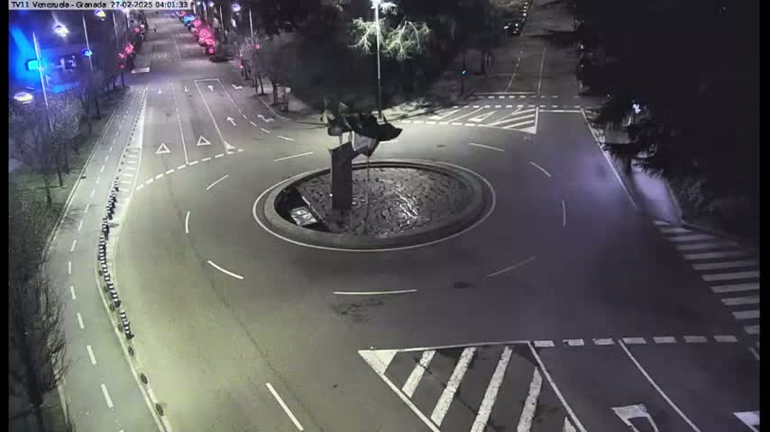 A nighttime aerial view shows a roundabout with a dark sculpture in the center, surrounded by roads with minimal traffic.