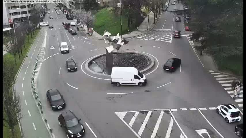 Several cars navigate a roundabout in a city, under a partly sunny sky.