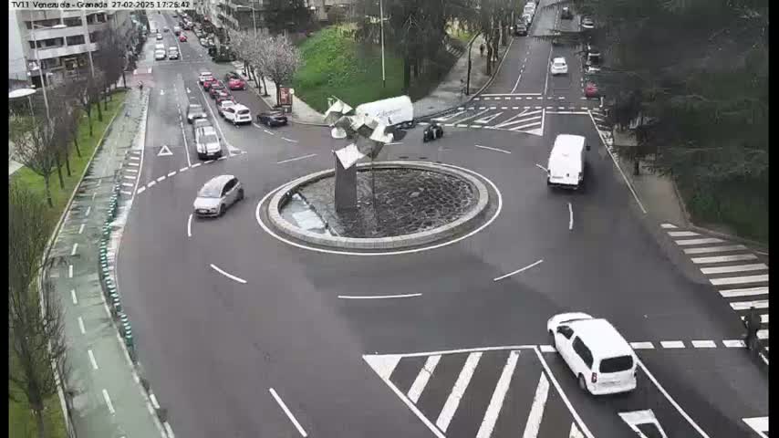 Several cars are driving around a roundabout featuring a modern sculpture in the center, on a partly cloudy day.