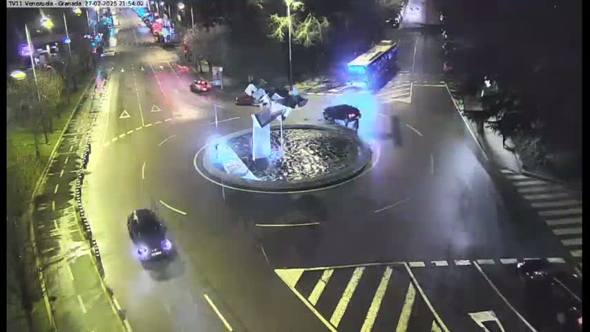 A nighttime aerial view shows a roundabout with several cars and a bus, and a fountain in the center, with wet roads suggesting recent rain.