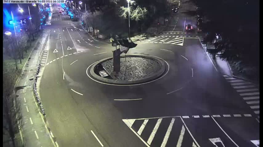 A nighttime aerial view shows a roundabout with a dark sculpture in the center, surrounded by wet roads and street markings.