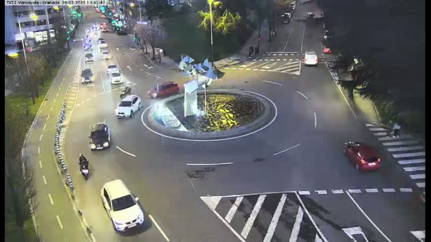 At night, several cars navigate a roundabout with a central sculpture, while a motorcycle and pedestrian are also visible near a crosswalk.