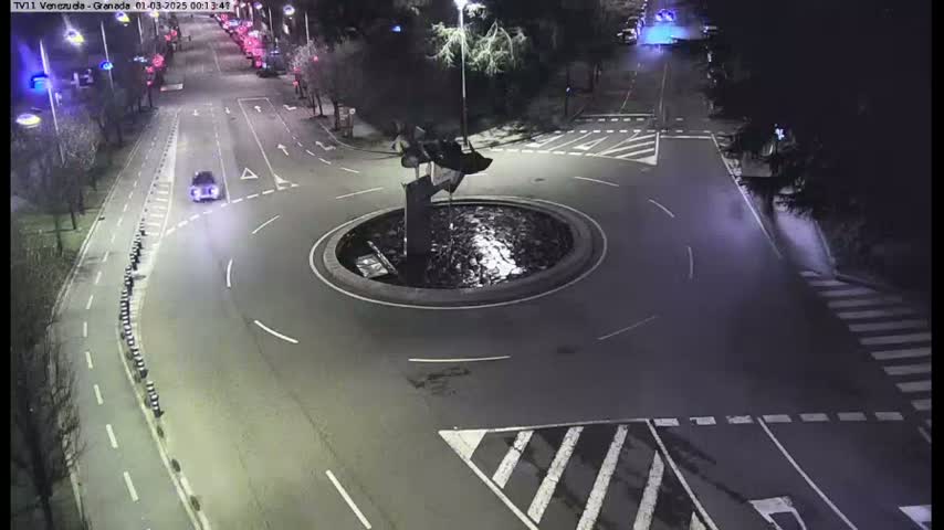 A nighttime aerial view shows a single car navigating a roundabout with a sculpture in the center, surrounded by streets and pedestrian crossings.