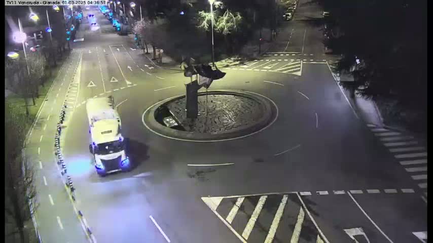 A large truck is navigating a roundabout at night, with a statue at its center.