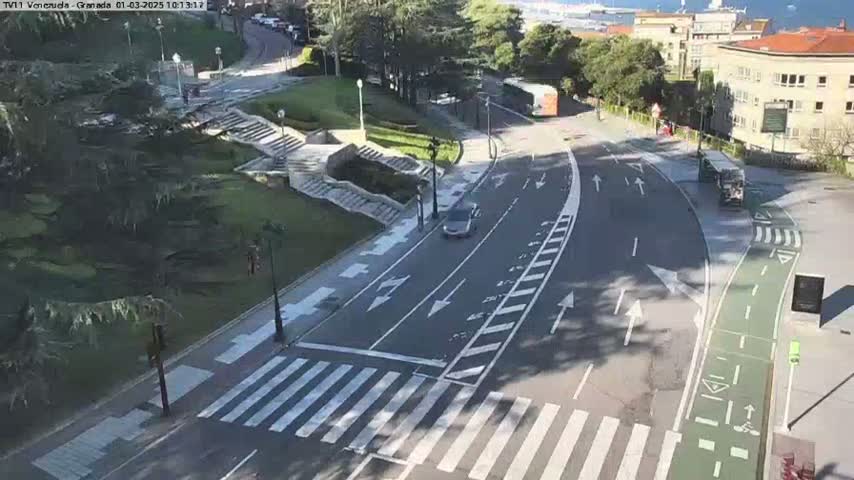 A sunny day overlooks a roadway with a car and a bus, marked crosswalks, and a bike lane next to a park with stairs and trees.