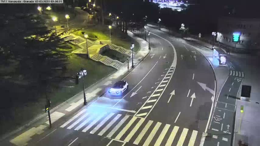 A white car drives on a curving road at night, past a crosswalk and stairs leading to a park-like area under illuminated streetlights.