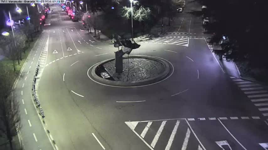 A nighttime aerial view of a roundabout with a dark-colored sculpture in the center, surrounded by roadways and pedestrian crossings.