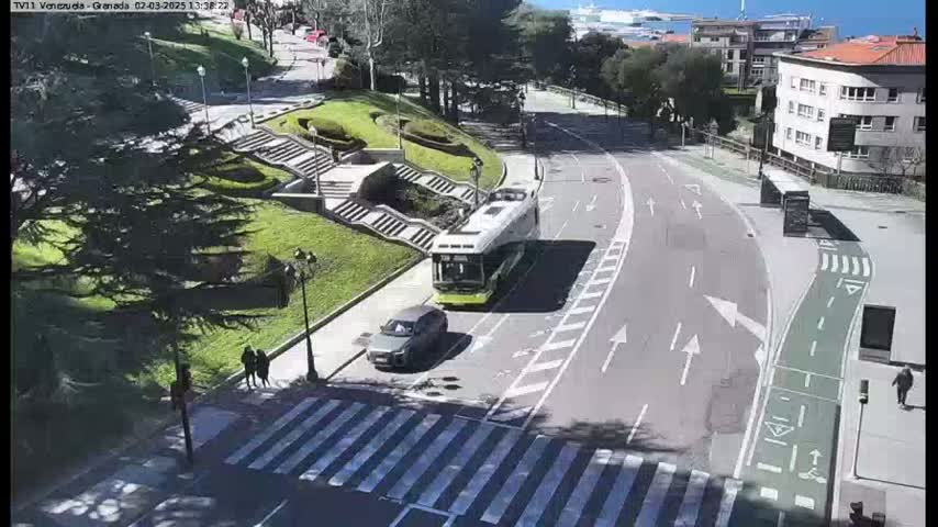 A city street scene on a sunny day shows a bus and car on a road that curves past a pedestrian crossing near stairs leading up a grassy hillside.