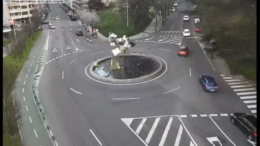 A roundabout with a modern sculpture in the center, surrounded by roads with several cars, under a partly cloudy sky.