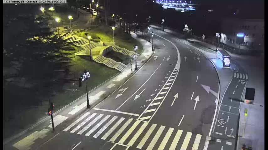 A nighttime, well-lit road curves through a park-like setting with stairs and landscaping, featuring a pedestrian crossing.