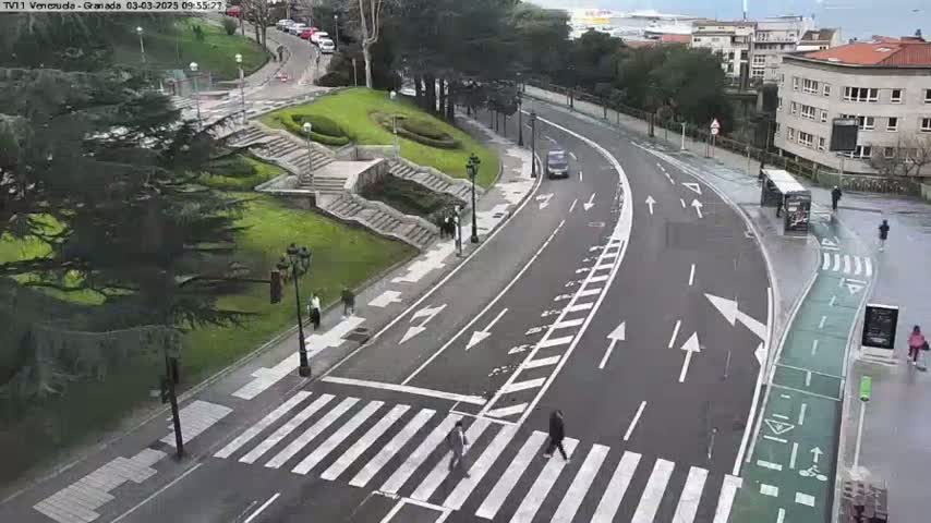 A paved road with crosswalks and bike lanes curves through a park-like area with stairs and greenery under an overcast sky.
