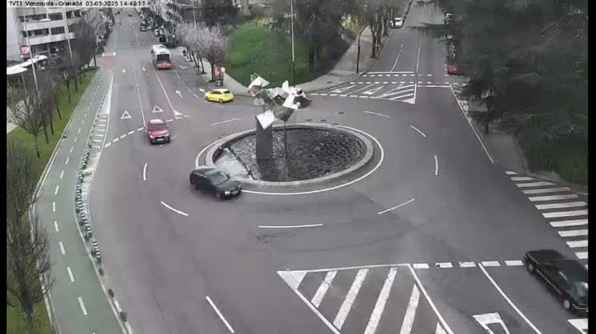 A traffic circle with a modern sculpture in the center is surrounded by several cars on a partly cloudy day.