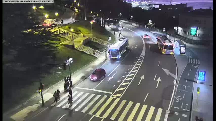 A nighttime street scene shows two buses and a car on a curved road with pedestrians crossing a crosswalk, near stairs and greenery.
