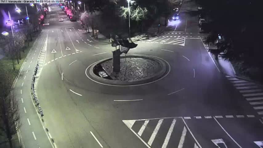 A nighttime aerial view of a roundabout with a dark sculpture in the center, surrounded by roads and pedestrian crossings.