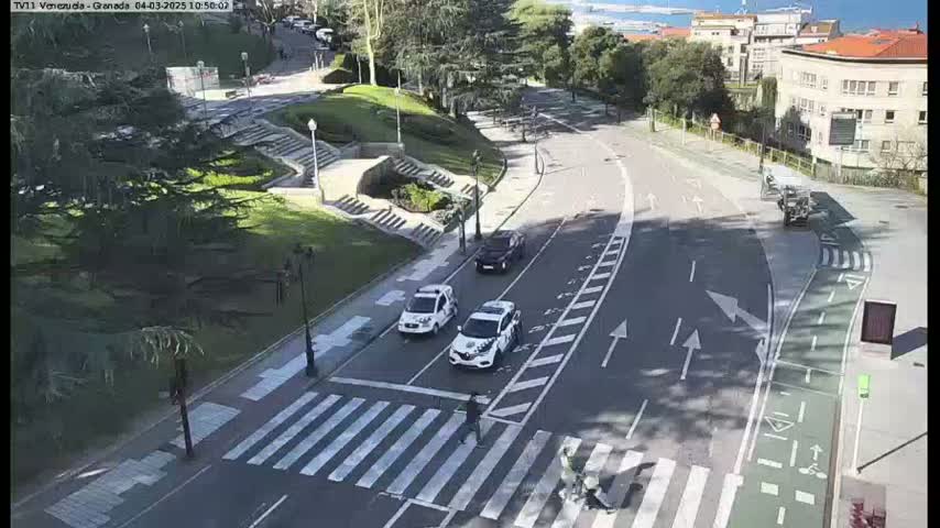 A sunny day shows a roadway curving through a city park with several cars and pedestrians crossing at a crosswalk.