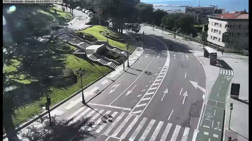 A sunny day overlooking a curving road with a crosswalk, bike lane, and stairs leading up to a grassy hill with buildings and a body of water visible in the distance.