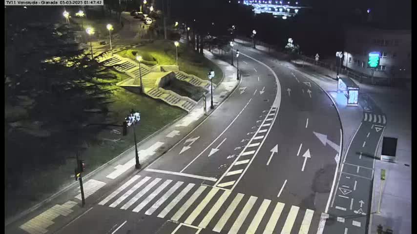 A nighttime view of a curved road with pedestrian crosswalks, stairs leading up to a grassy area, and streetlights illuminating the scene.
