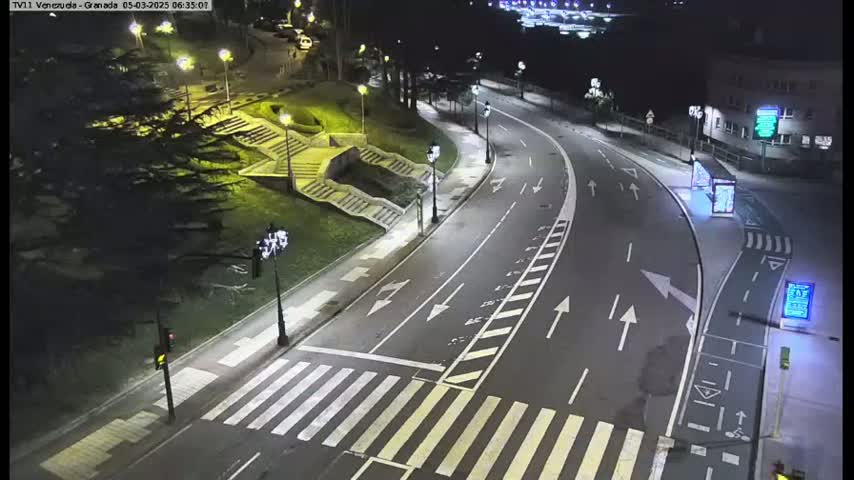 A nighttime view of a mostly empty road curving through a park-like area with stairs and landscaping, under artificial light.