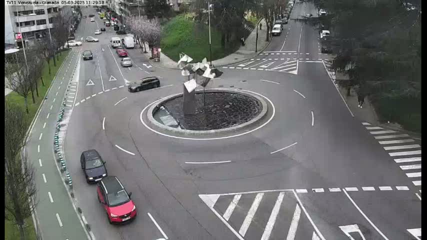 A roundabout with several cars and a modern metal sculpture in the center is situated at the intersection of several roads, on an overcast day.