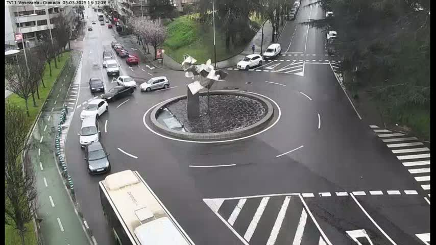 A wet roundabout with several cars and a bus is surrounded by roads and a pedestrian crossing, with trees and buildings visible in the background.