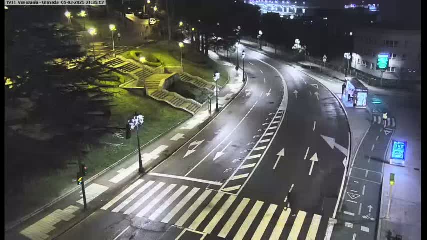 A nighttime, high-angle view of a mostly empty road curving through a landscaped area with stairs and a pedestrian crossing, illuminated by streetlights.