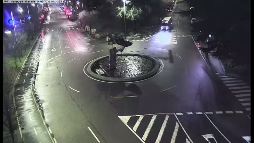 A nighttime aerial view shows a roundabout with a dark sculpture in the center, surrounded by streets and crosswalks, with a few vehicles visible.