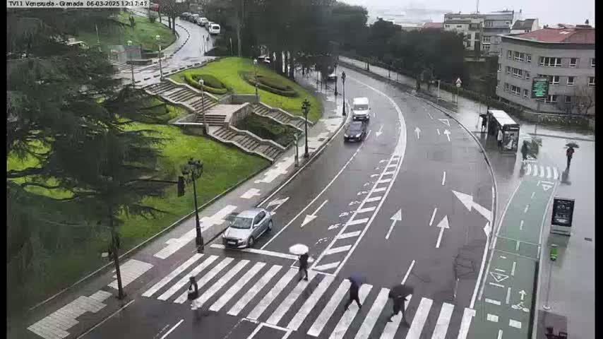 A rain-slicked road curves past a pedestrian crossing and stairs leading up to a grassy hill with trees, with several people walking under umbrellas.