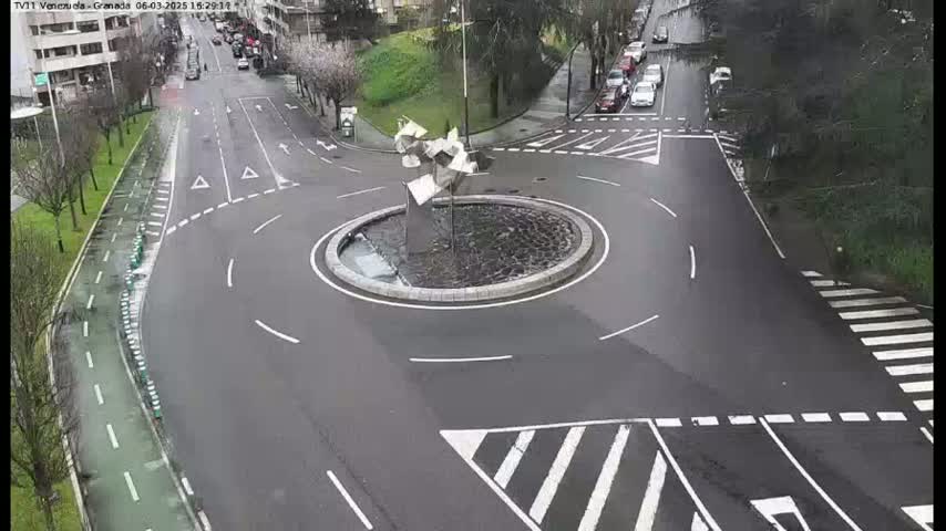A roundabout with a modern metal sculpture in the center is surrounded by wet roads and pedestrian crossings on a cloudy day.