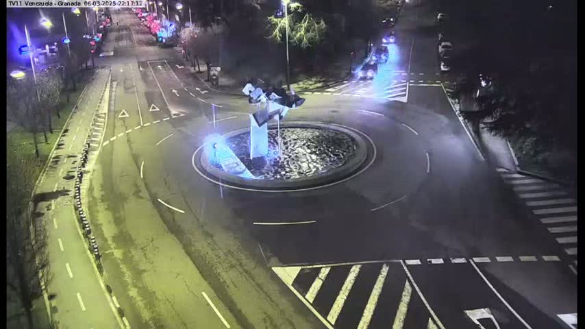 A nighttime aerial view shows a roundabout with a modern sculpture in the center, surrounded by roads with sparse traffic.