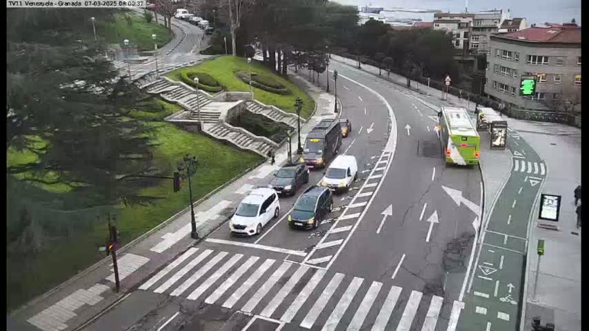 Several cars and a bus are on a roadway curving around a grassy area with stairs, under an overcast sky.
