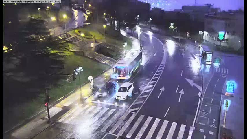 A rainy night scene shows a crosswalk with several cars and a bus at an intersection, with wet roads and streetlights illuminating the area.