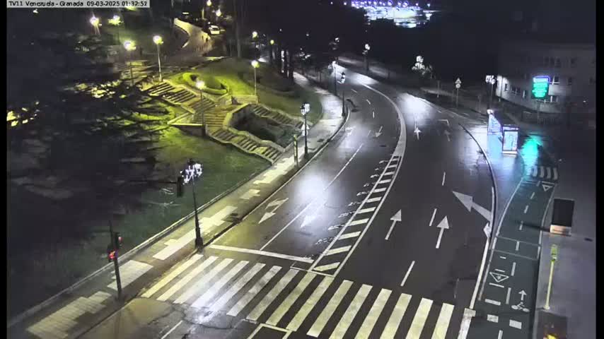 A wet, nighttime street scene shows a curving road with crosswalks and a bike lane, leading past stairs and landscaping toward a body of water.