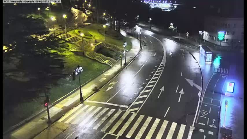 A wet, empty roadway curves through a city at night, illuminated by streetlights, with pedestrian crosswalks and stairs visible on the side.