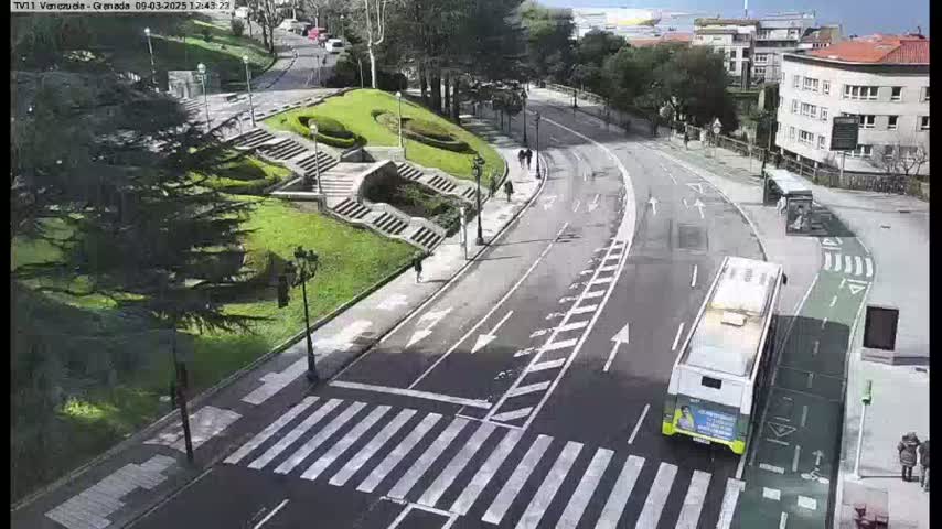 A sunny day shows a curving road with pedestrian crosswalks and a bus, passing by a grassy area with stairs and trees.