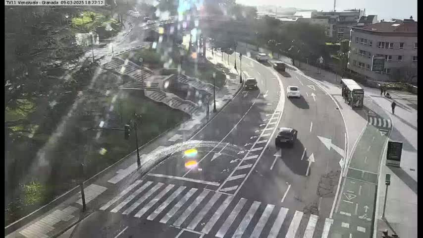 A rainy day scene shows a road intersection with several cars and a bus, a pedestrian crossing, and a bike lane, all near a set of stairs leading down to a park-like area with trees.