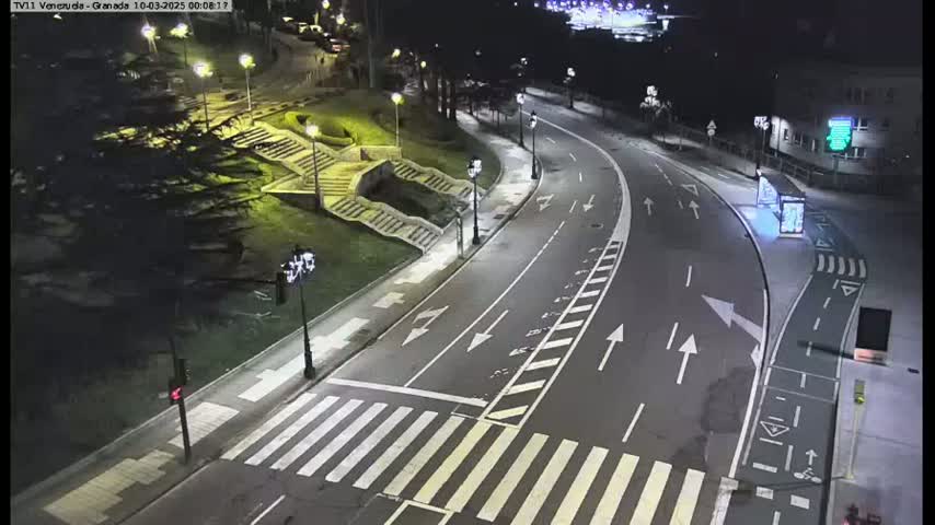 A nighttime, brightly lit road curves through a park-like setting with stairs and landscaping, featuring a pedestrian crossing.