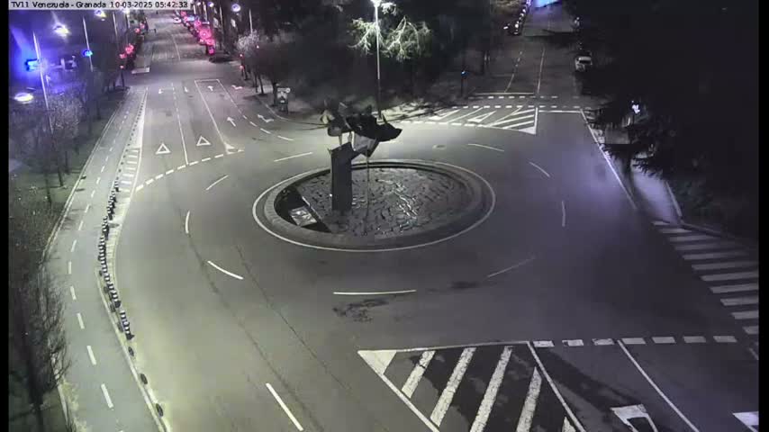 A nighttime aerial view shows a roundabout with a dark sculpture in the center, surrounded by roads and pedestrian crossings.