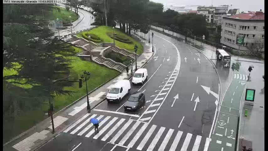 A rainy street scene shows a pedestrian crossing a crosswalk, several cars parked and driving on a curved road next to a grassy area with stairs, and a bus stop.
