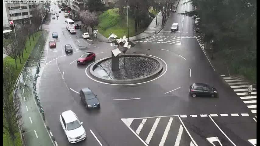 A wet roundabout with several cars and a metal sculpture in the center is viewed from above.