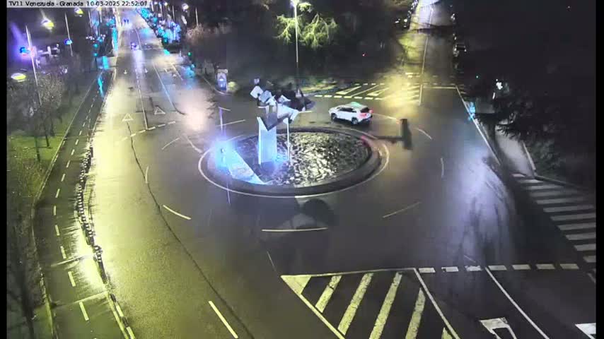 A wet, nighttime street scene shows a roundabout with a modern sculpture in the center, with a car visible on the roundabout and other vehicles on the road.
