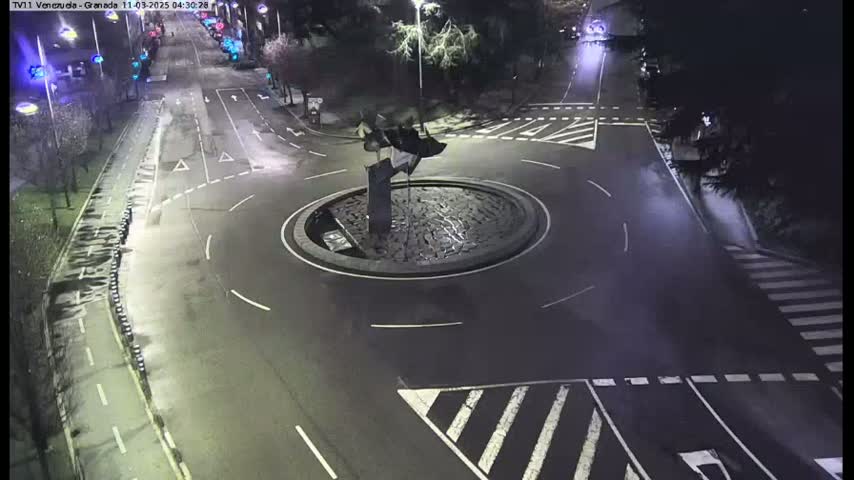 A nighttime aerial view shows a roundabout with a dark sculpture in the center, surrounded by wet asphalt roads and pedestrian crossings.